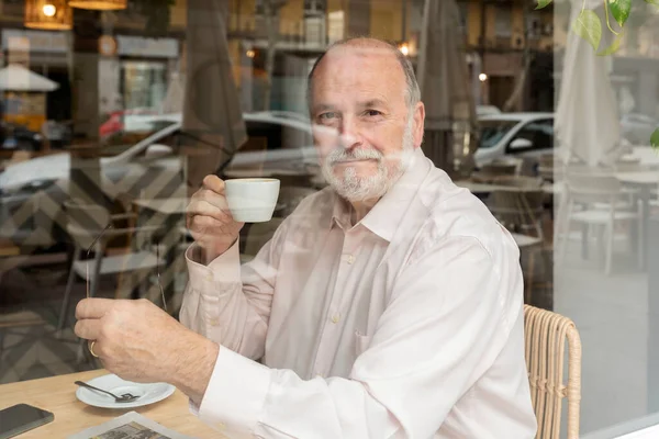 Kahve barında oturan yaşlı bir adam elinde kahve fincanı ve gözlükleriyle pencereden kameraya bakıyor.