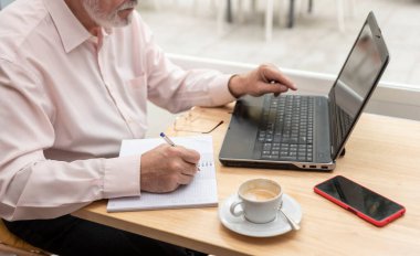 Önünde dizüstü bilgisayarıyla masada oturan tanınmayan yaşlı bir adam defterine, kahve fincanına ve yanındaki cep telefonuna finansal notlar alıyor.