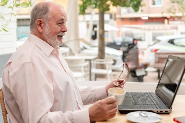 Yaşlı bir adam kahve dükkanında oturuyor, gözlüğünü tutuyor, gülüyor ve dizüstü bilgisayarına bakıyor.