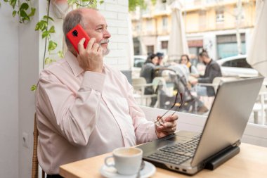 Kahve dükkanında gülümseyen yaşlı bir adam camdan dışarı bakıyor. Gözlüğünü tutuyor ve cep telefonuyla konuşuyor. Dizüstü bilgisayarında çalışmaya ara veriyor.