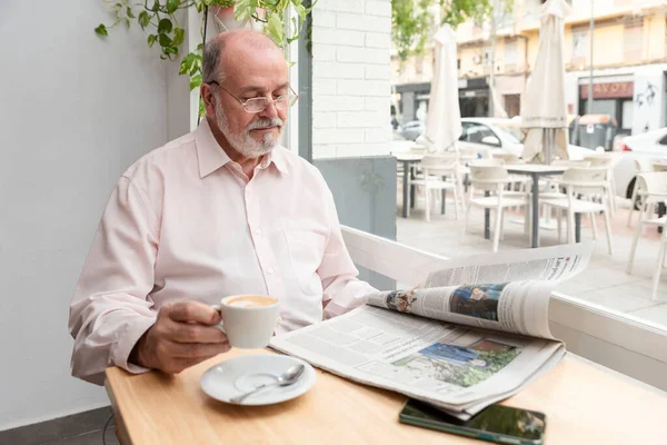 Gözlüklü yaşlı bir adam kahve dükkanında oturmuş günlük gazete okuyor. Elinde bir fincan kahve, yanında cep telefonu, ahşap masanın üstünde.