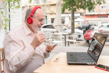 Kulaklıklı gülümseyen yaşlı bir adam kahve fincanının tadını çıkarıyor. Dil öğrenmek için dizüstü bilgisayar kullanıyor, online dersler veriyor, emekliliğinin tadını çıkarıyor.