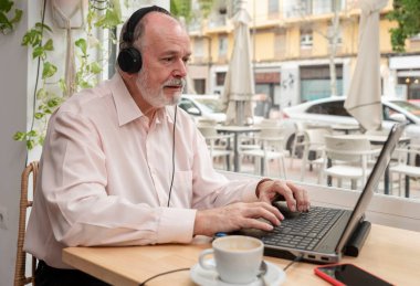 Yaşlı bir adam, bir kafede oturmuş, kulaklıklarıyla bilgisayarını kullanarak internetten ders izliyor ve kahvenin tadını çıkarıyordu.