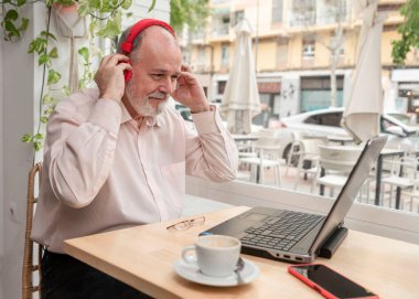 Kahve dükkanında gülümseyen yaşlı bir adam kahvesini içerken müzik dinlemek için dizüstü bilgisayar kullanır.