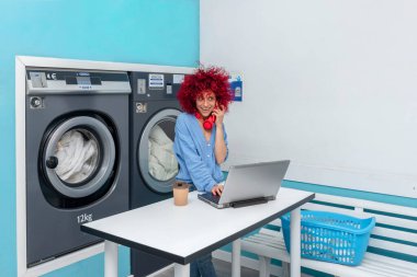 Kırmızı saçlı, gülümseyen bir Latin kadın laptopuyla çalışıyor ve mavi çamaşır odasında telefonla konuşuyor. Çamaşırların yıkanmasını bekliyor. Kırmızı kulaklıkları boynunda.