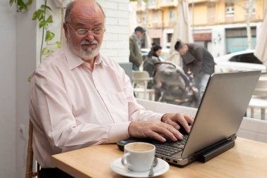 Kafede dizüstü bilgisayarıyla oturan yaşlı bir adam komik bir yüz ifadesi takınıyor.