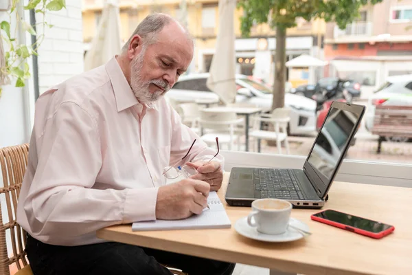 an older man sitting at a table with his laptop in front and taking notes of his business finances in his notebook, coffee cup and mobile phone next to him