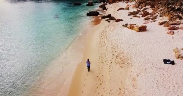 Sportowa blondynka biegająca po pustej plaży. Kobieta ćwiczy na świeżym powietrzu biegając po skalistej plaży. Koncepcja zdrowego biegania i ćwiczeń na świeżym powietrzu. Aktywny, sportowy jogging. Dron — Wideo stockowe