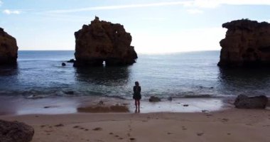 Boş Algarve sahilinde yürüyen güzel el sallayan elbiseli bir kadın. Drone havalanıyor ve kamerayla yukarı çıkıyor. Kristal berrak su ve kayalık sahilde yürüyorum. 