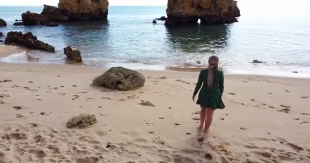 Camera slowly tilting up while a beautiful woman is walking to the beach. Calm waves and rocky coastline. Colorful golden beach. Happy beautiful women. Crystal clear water and water reflecting. — Stock Video