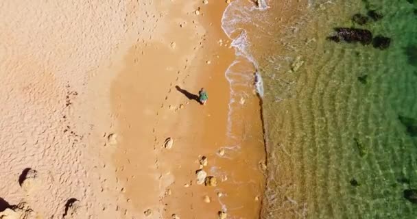Kamera podąża za kobietą, która idzie samotnie po plaży, kamera powoli pochyla się nad skalistym wybrzeżem. Kolorowa złota plaża. Szczęśliwe piękne kobiety. Krystalicznie czysta woda i woda odbijające — Wideo stockowe