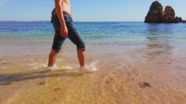 Male legs walking along the sea coast. Slowed down walking at the beach. Vacation at the beach. Small waves hitting their feet on the shore. Stone archs in the ocean at the background — стоковое видео
