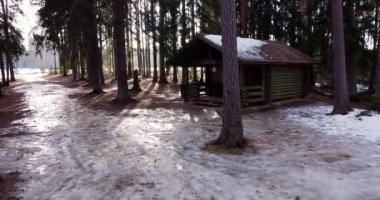 Flying through forest overlooking log cabin covered with snow. Sun is shining through trees. Sun rays through trees. Beautiful scenery view. Little cozy house in the forest. Wooden cabin. Winter time
