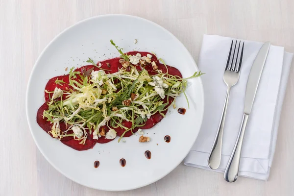 Beetroot carpaccio with arugula, dor blue, walnuts and salad on a plate. A plate of vegan red beetroot carpaccio on the table with a knife and a spoon.