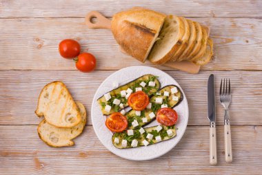 Beyaz ahşap zemin üzerinde peynirli ızgara patlıcan ve ızgara domates dilimlenmiş ekmek ve kızarmış ekmek, sarımsak, çatal ve bıçak. Yukarıdan bak. Tabakta ızgara sebze..