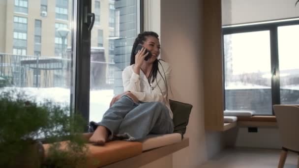 Egy gyönyörű fiatal lány ül az ablakpárkányon az ablaknál, és telefonon beszél a szeretteivel. Kommunikációs elv. — Stock videók