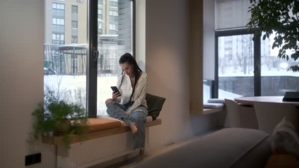 Una bella ragazza si siede sul davanzale della finestra e parla al telefono con i propri cari. Concetto di comunicazione. — Video Stock