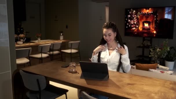 A beautiful young girl with dreadlocks sits at a table with a tablet and communicates via video link with her family. Communication concept. — Stock Video