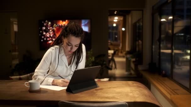 En flicka med dreadlocks i en vit skjorta studier med hjälp av en digital tablett i en online-lektion, skriver ner de viktiga punkterna i lektionen. Begreppet lärande online. — Stockvideo