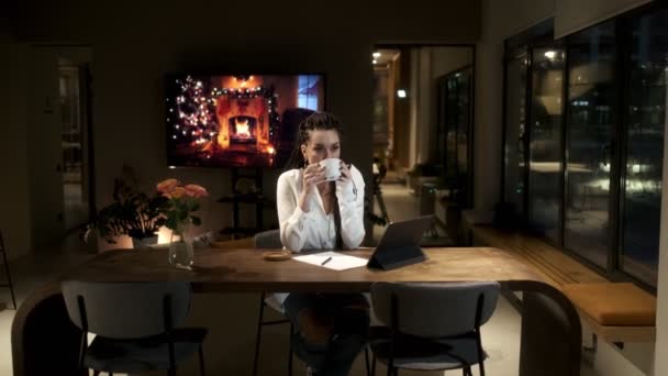 En pige med dreadlocks i en hvid skjorte drikker kaffe og studerer på en tablet i en online lektion. Pause fra klassen. Online læringskoncept. – Stock-video