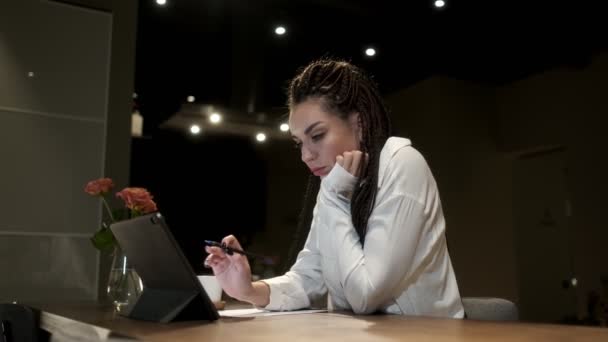 Een meisje met dreadlocks in een wit shirt bestudeert artikelen op een tablet voor haar werk. Online leerconcept. — Stockvideo