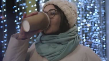 Gözlüklü güzel bir esmer ve gece şehrinde bir kış şapkası soğuk gölgeli bir çelengin arka planına karşı kahve içer ve ısınır. Bekleme ve keyif konsepti.