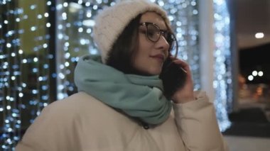 Kışın gözlüklü genç bir kız telefonda çelenk duvarının arkasında konuşuyor. İletişim konsepti.