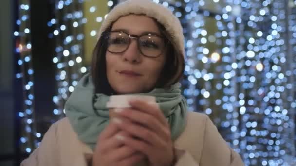 Eine schöne Brünette in Brille und Wintermütze in der nächtlichen Stadt vor dem Hintergrund einer Girlande aus kaltem Schatten trinkt Kaffee und hält sich warm. Warten und Freude. — Stockvideo