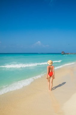 Kırmızı mayo giymiş genç bir kadın sırtı sahilde duruyor.