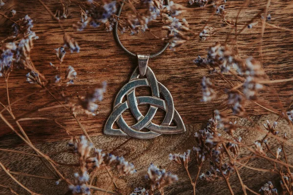 Kuru çiçeklerdeki ahşap bir masada gümüş bir triquetra kolye.
