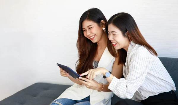 两个年轻的亚洲女孩穿着办公室风格的衣服 坐在沙发上 使用平板电脑 带着快乐 欢乐和笑脸为互联网服务 几个女同事在网上购物 — 图库照片