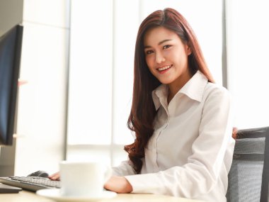 A beautiful, friendly, and self-assured corporate woman sits at her office desk in the orange light of the morning sun. Smile brightly and look at the camera.