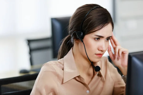 A beautiful woman in a headphone call center worker working at a computer screen stressing her face during a conversation with a customer.