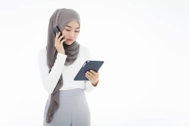 A beautiful Asian Muslim woman wearing a hijab, stylish and tech-savvy, is using smartphones and tablet computers to work and communicate. Islamic woman concept of doing business and communicating