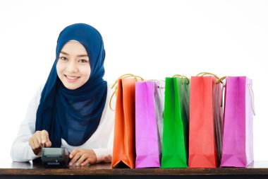 A young, modern-looking Asian Muslim woman is using a credit card reader. Islamic female shop owner making cash register beside colorful shopping bags. Buying concept and financial lifestyle.