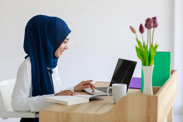 A beautiful young Asian Muslim woman wearing a beautiful hijab and headscarf is researching with a laptop computer and a textbook. Concept of belief and learning.