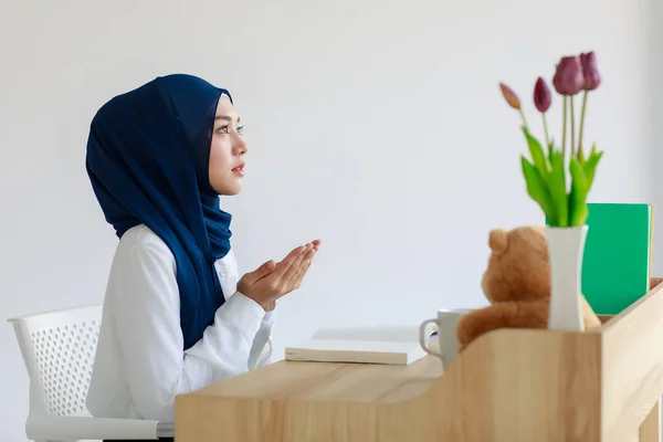 A beautiful young Asian Muslim woman wearing a beautiful hijab and headscarf is raising her hands in prayer. A female Islamic student prays while reading a book. Concept of belief and learning.