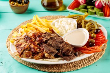 Türk Karışık Shawarma Tabağı, patates kızartması, mayonez sosu, sos ve salata. Tahta kenarından kesilmiş bir tabakta servis ediliyor.
