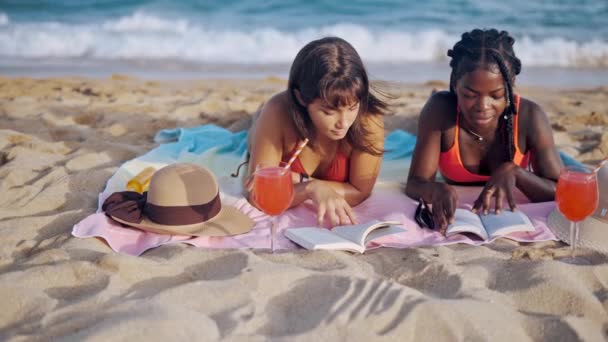 Дві дівчини читають книги, лежачи на піщаному узбережжі — стокове відео