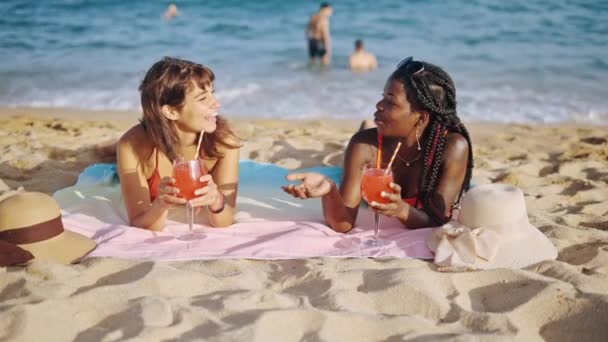 Due giovani ragazze in costume da bagno sdraiate sulla spiaggia di sabbia con cocktail di frutta, parlando e sorridendo — Video Stock