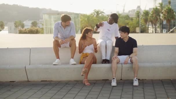 Four young multi-ethnic friends sitting on concrete bench outdoors at sunset and communicating — Stock Video