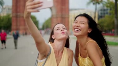 İki mutlu melez kadın arkadaş dışarıda selfie çekiyorlar ve cep telefonundaki en iyi fotoğrafı seçiyorlar.