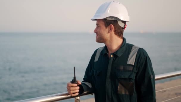Šťastný přístavní dělník s vysílačkou v ruce stojící v přístavním terminálu, užívající si výhled na moře — Stock video