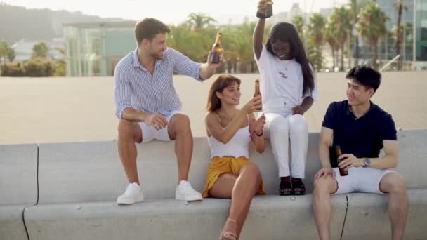 Улыбающиеся разнообразные друзья, звонящие бутылками, когда пьют пиво на закате на набережной в выходные — стоковое видео