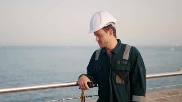 Young man port worker in helmet and coverall looking at sea while standing at dockside — Stock Video