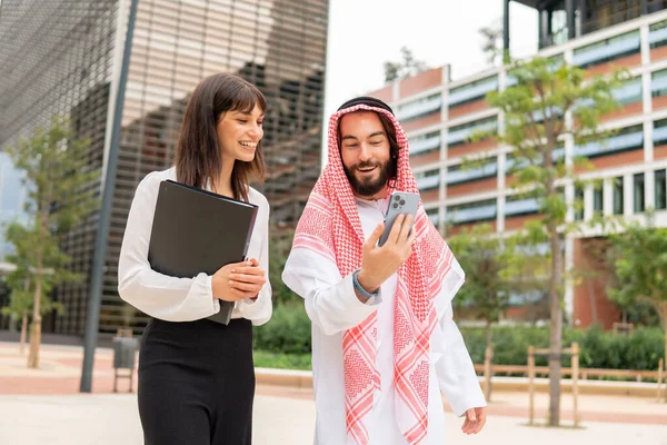 Smiling arab businessman and young caucasian businesswoman using smartphone at meeting outdoors — Stock Photo, Image