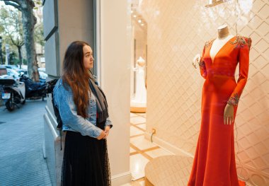 Şehirdeki moda mağazasının vitrininin arkasına yerleştirilmiş lüks kırmızı elbiseyi beğenen rüya gibi bir kadının yan görüntüsü.
