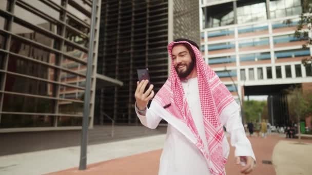 Tradicional vestido hombre árabe feliz habla con la familia en línea a través de teléfono inteligente mientras camina calle de la ciudad — Vídeos de Stock