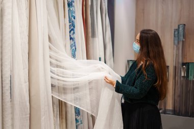 Yüz maskeli genç bir kadın kumaş mağazasında tül ve perde seçiyor, Covid19 sırasında alışveriş yapıyor.