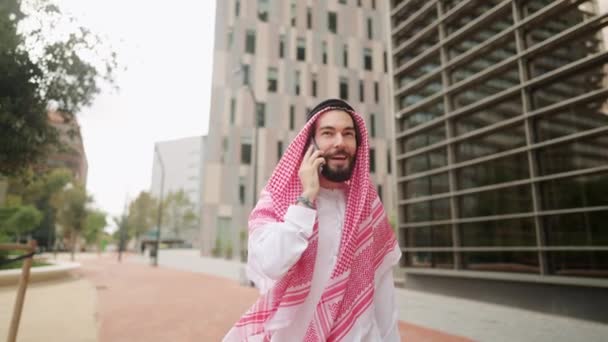 Smiling arab man enjoying phone conversation outdoors, walking city street in Dubai — Stock Video